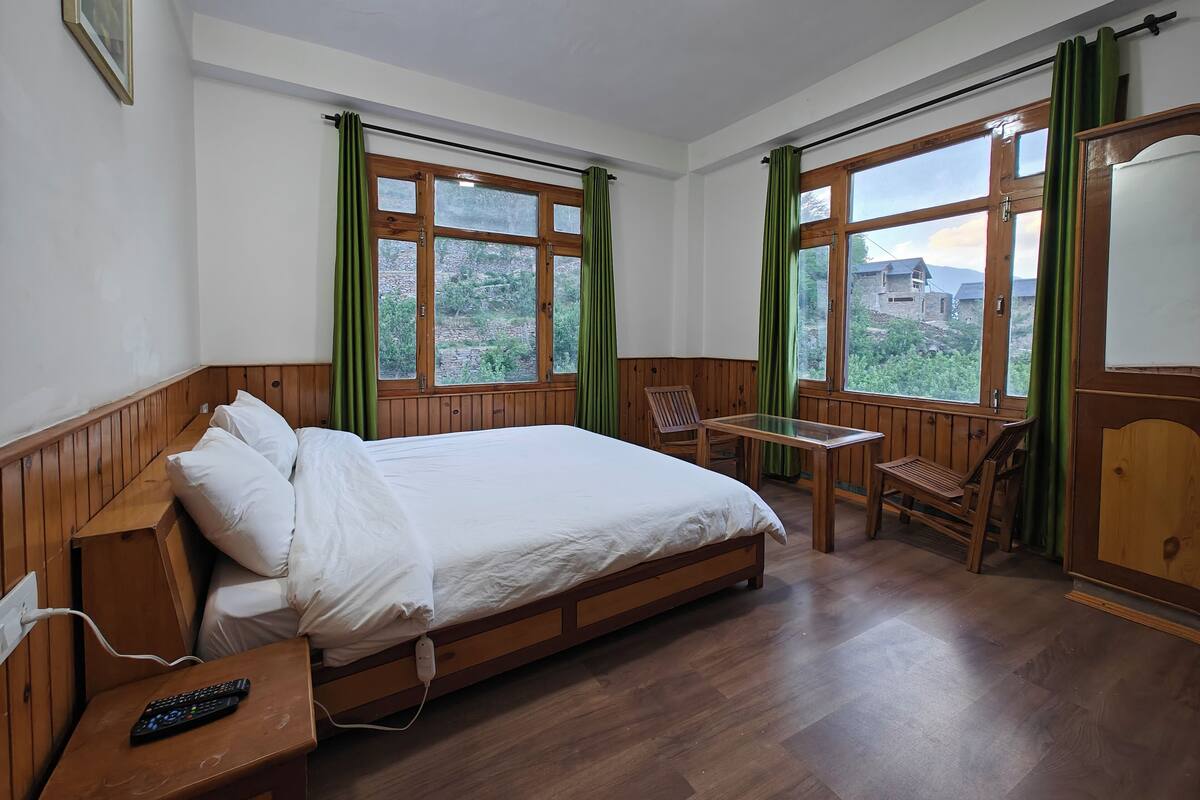 A bedroom with a wooden bed, a desk, and a chair, large windows with curtains, and a view of the outdoors.