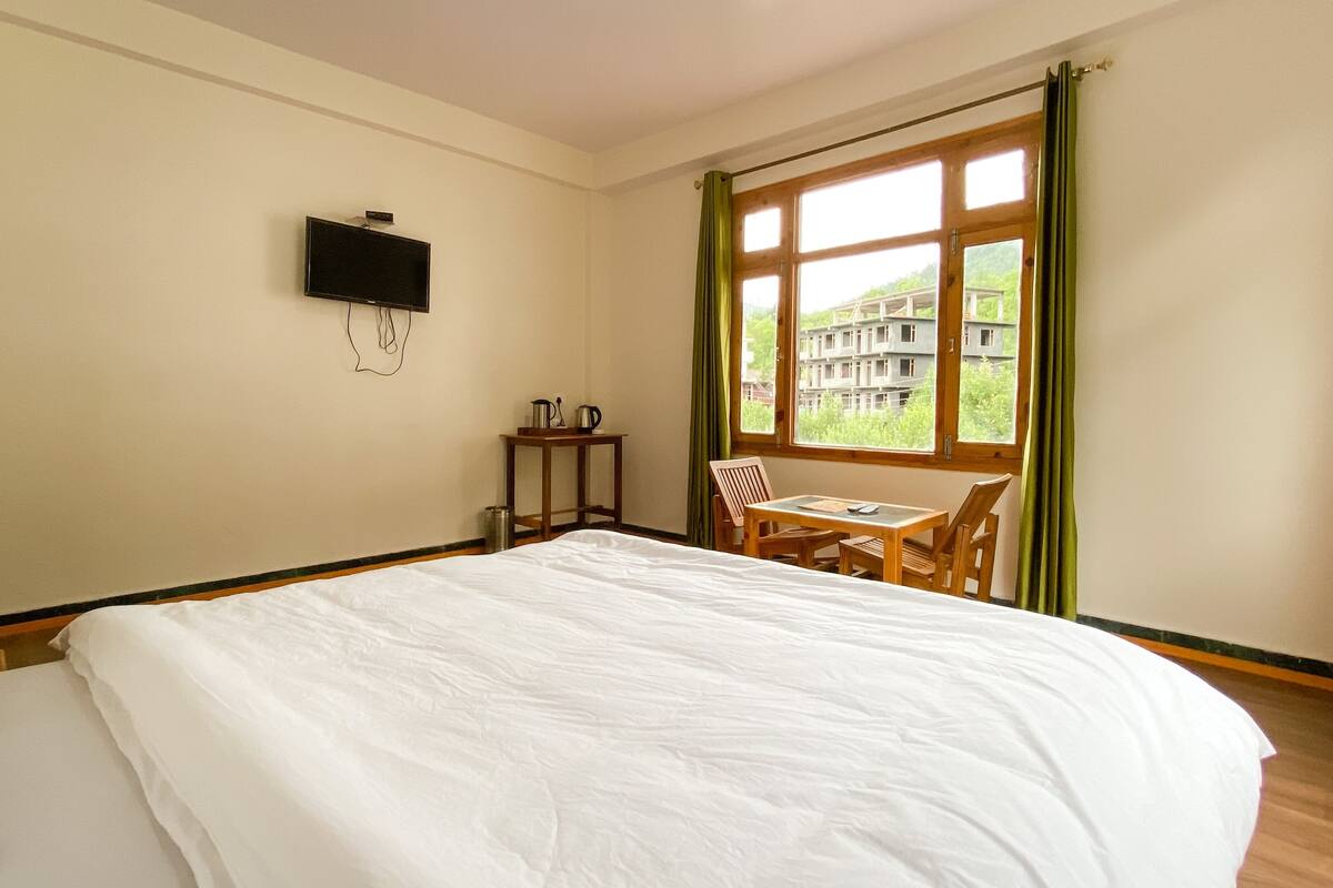 A hotel room with a bed, a small table with chairs, a television on the wall, and a window with curtains.