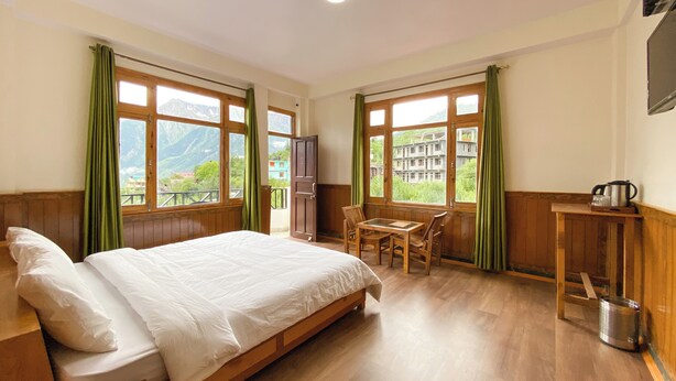 Une chambre d’hôtel avec un lit en bois, une petite table avec des chaises et une vue sur les montagnes, visible à travers de grandes fenêtres.