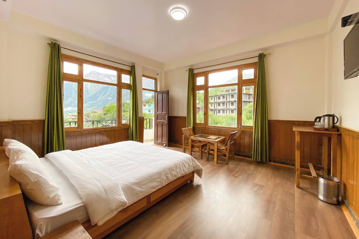 A hotel room with a wooden bed, a small table with chairs, and a view of mountains through large windows.