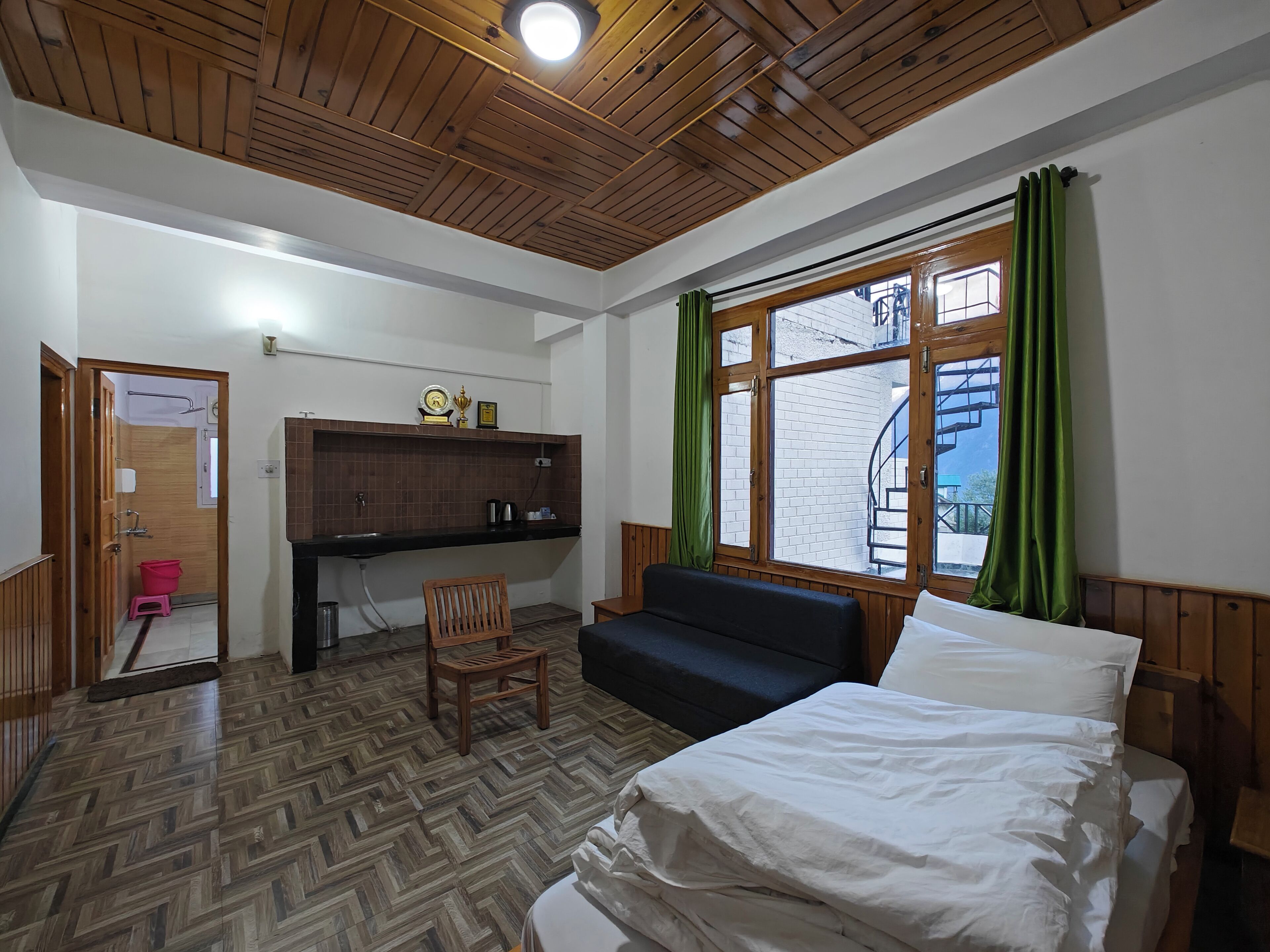 Une pièce avec un lit, un plafond en bois, une fenêtre avec des rideaux verts, un petit bureau avec une chaise, et une vue sur un bâtiment en briques à l’extérieur.