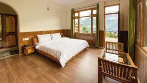 Une chambre avec un lit en bois, une table de chevet, une table en verre avec une chaise, et une vue sur les montagnes à travers de grandes fenêtres.