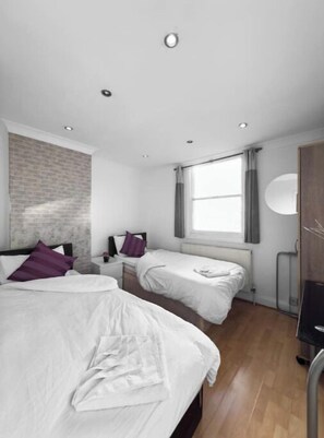 Two single beds with white linens and purple pillows, set parallel to each other, in a room with a textured brick pattern and a large window.
