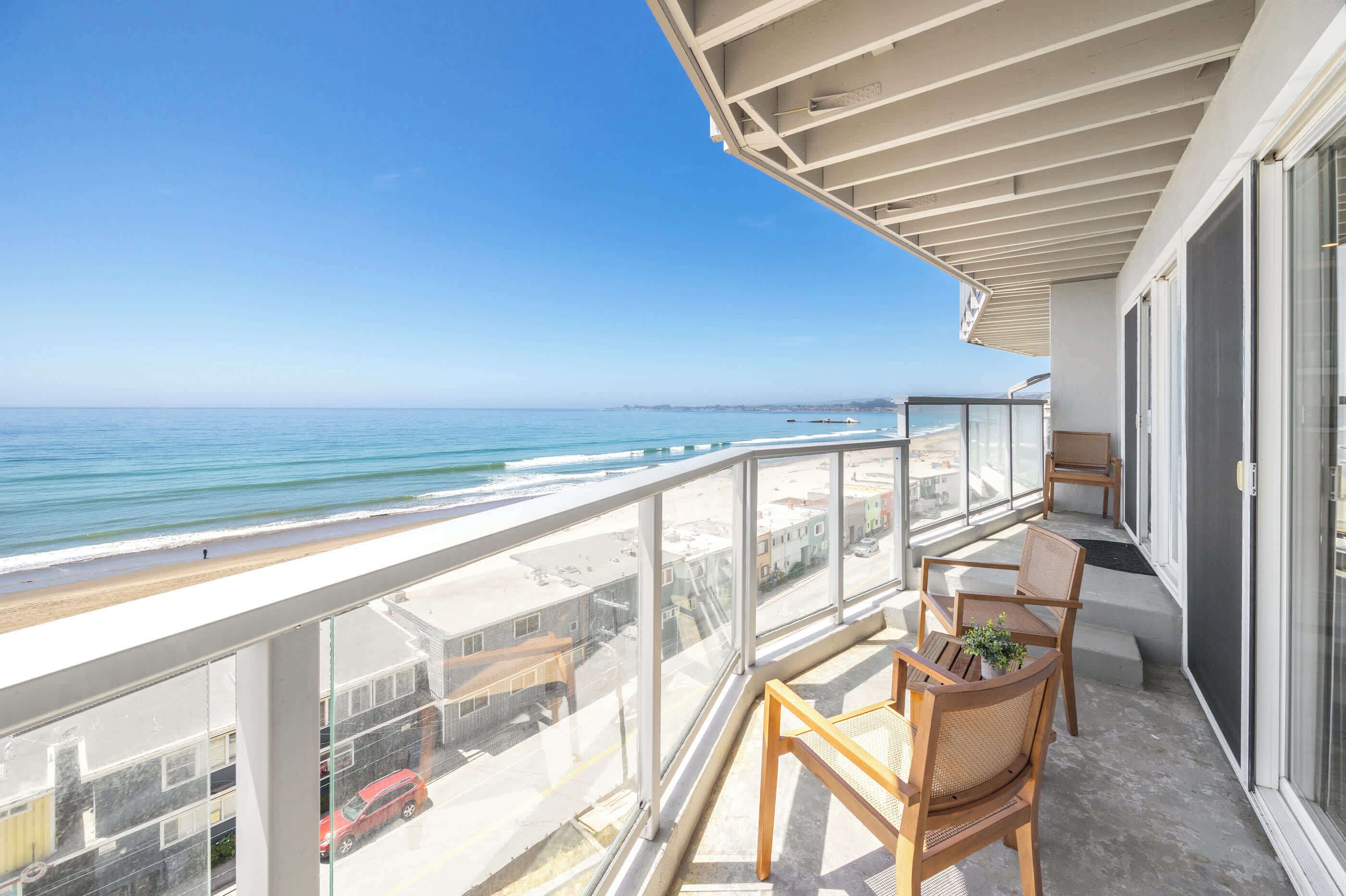 Un balcón con vista a la playa y al océano, sillas de madera y barandilla de vidrio.