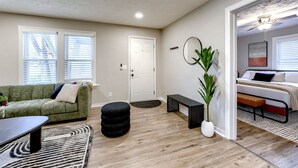 A living room with a green sofa, a black ottoman, a coffee table, and a patterned rug. There is a door leading to a bedroom with a bed and a ceiling fan. - Dundee Delight 3Br2Ba sleeps 9, central Omaha, heart of historical neighborhood (Omaha)