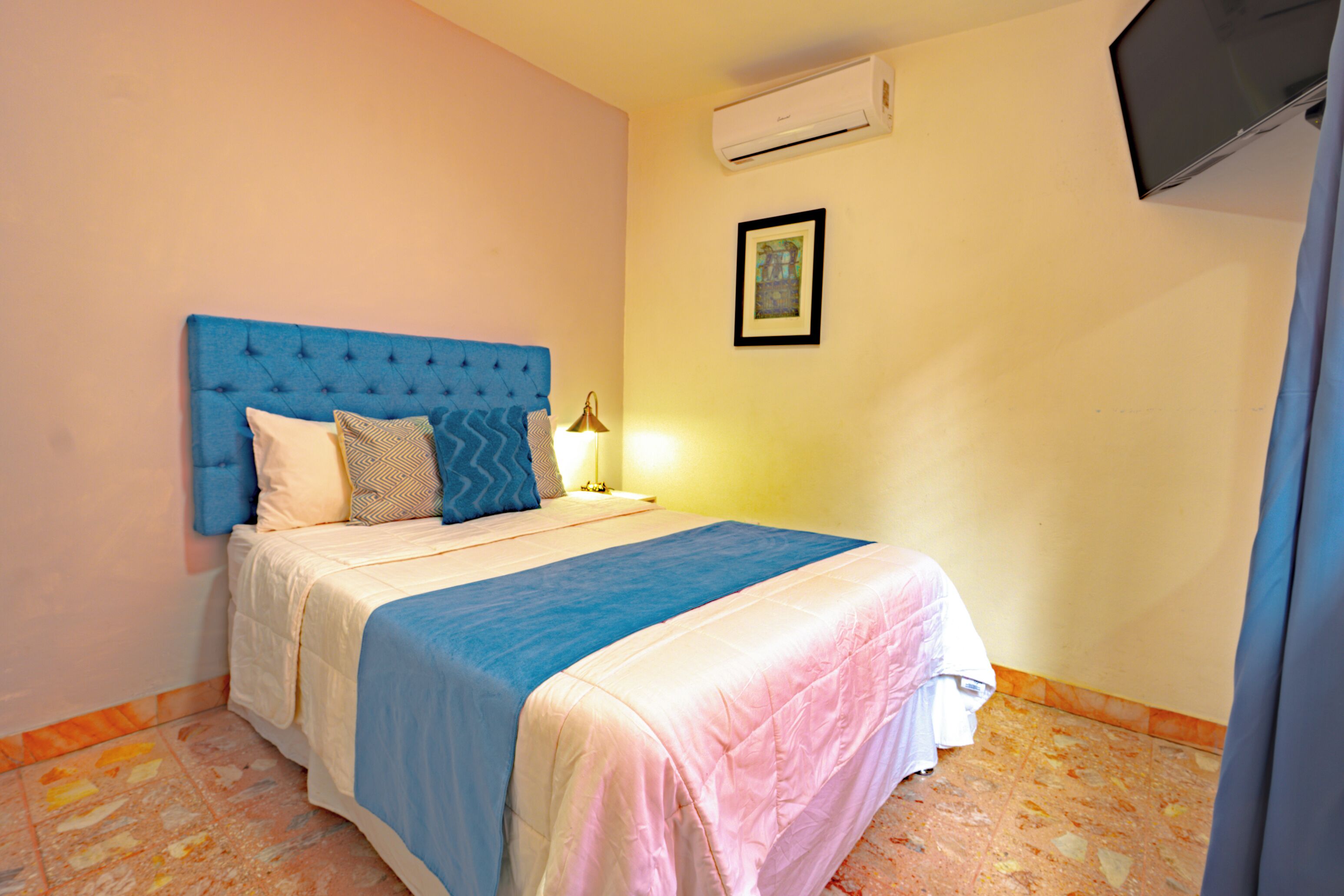 Ein Schlafzimmer mit blauem Kopfteil, einem Bett mit blauer und pinkfarbener Bettdecke, einer Nachttischlampe und einer Klimaanlage an der Wand.