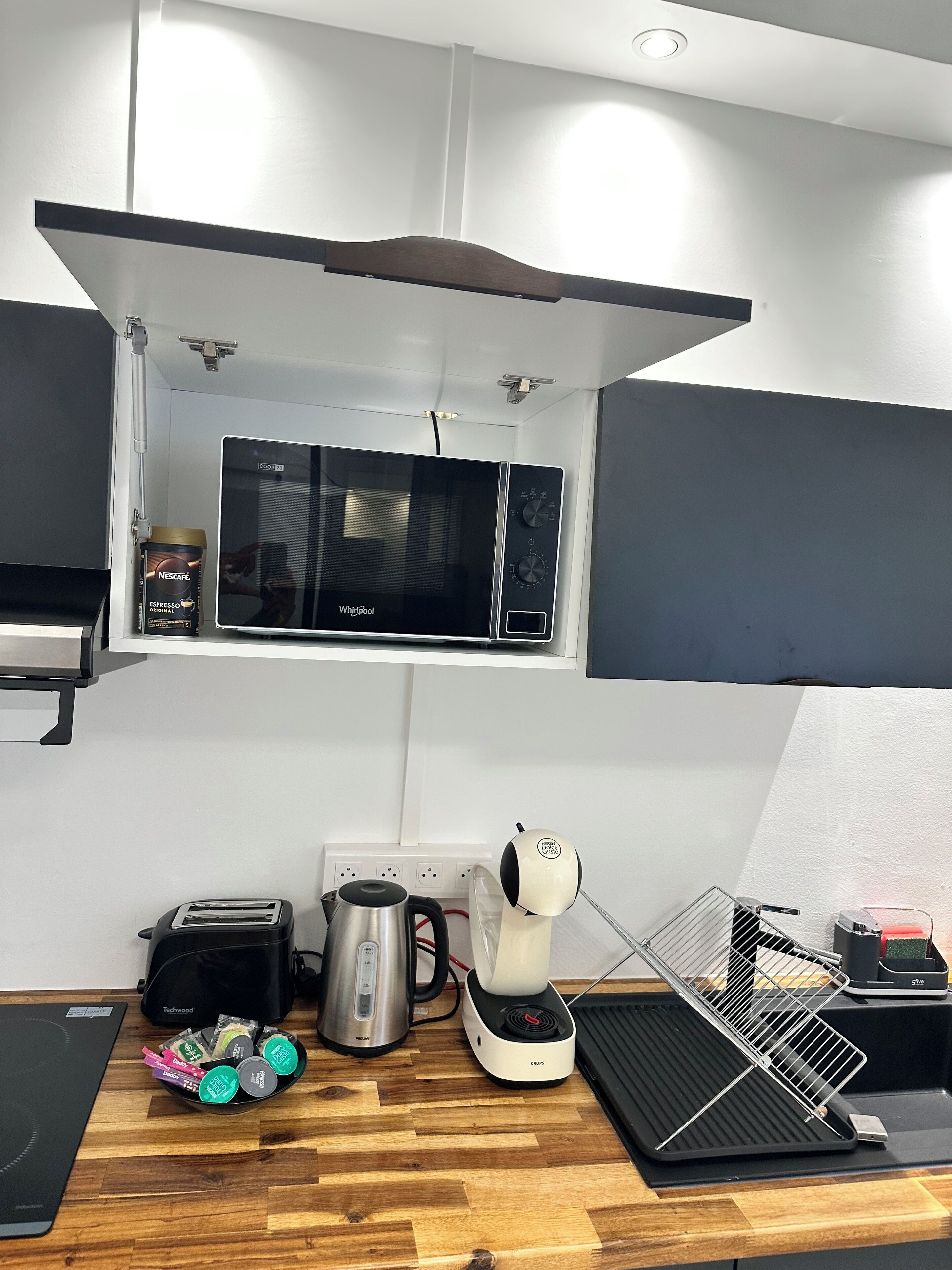 Cocina compacta con microondas, hervidor, tostadora y cafetera sobre una encimera de madera.