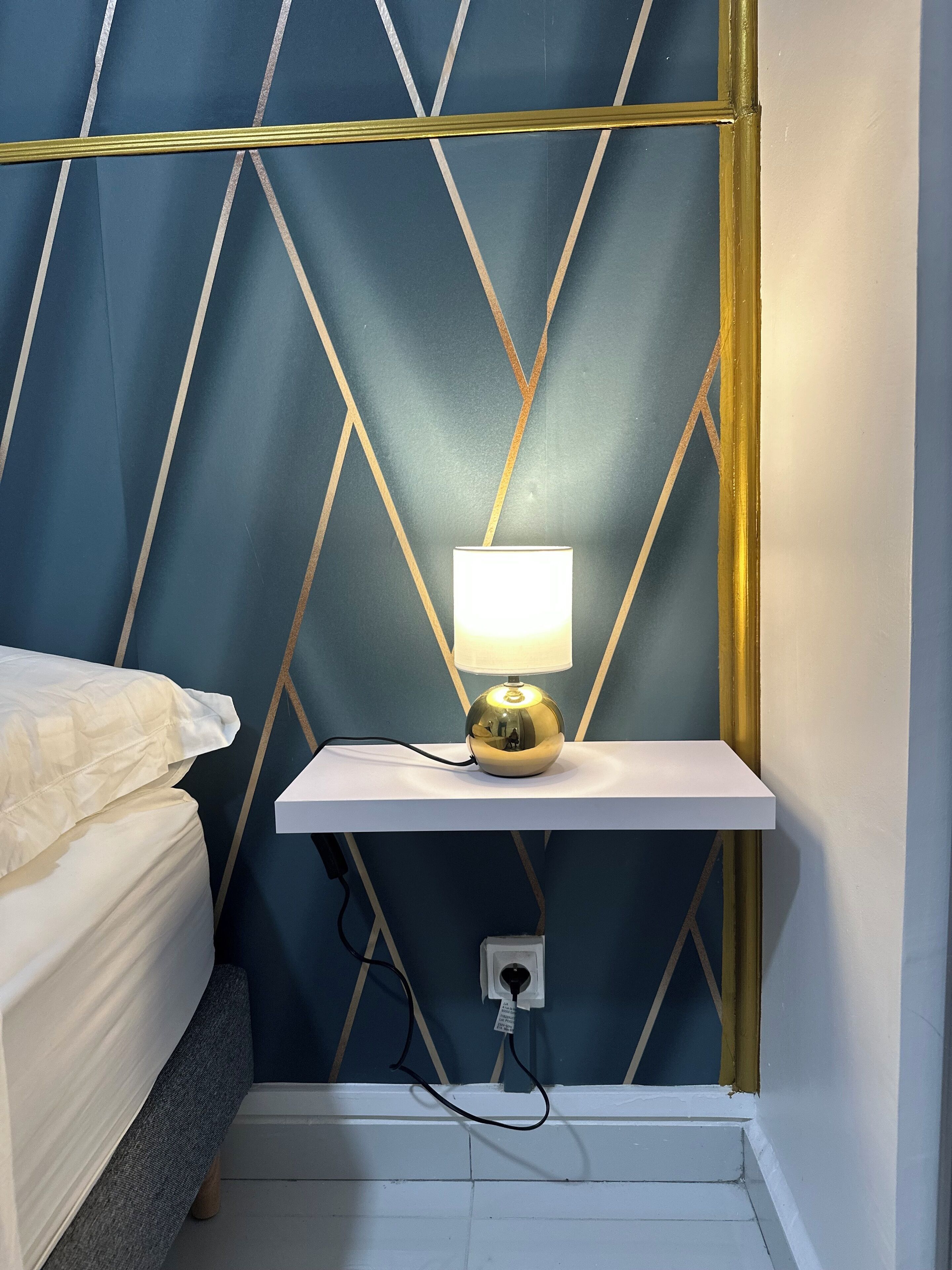 A bed with a white headboard, a bedside table with a lamp, and a wall with a geometric pattern.