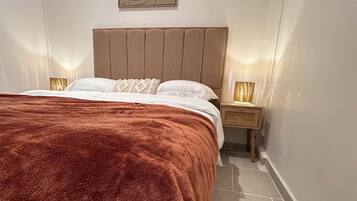 Un lit bien fait, avec une couverture marron, une tête de lit en bois et une table de chevet surmontée d’une lampe.