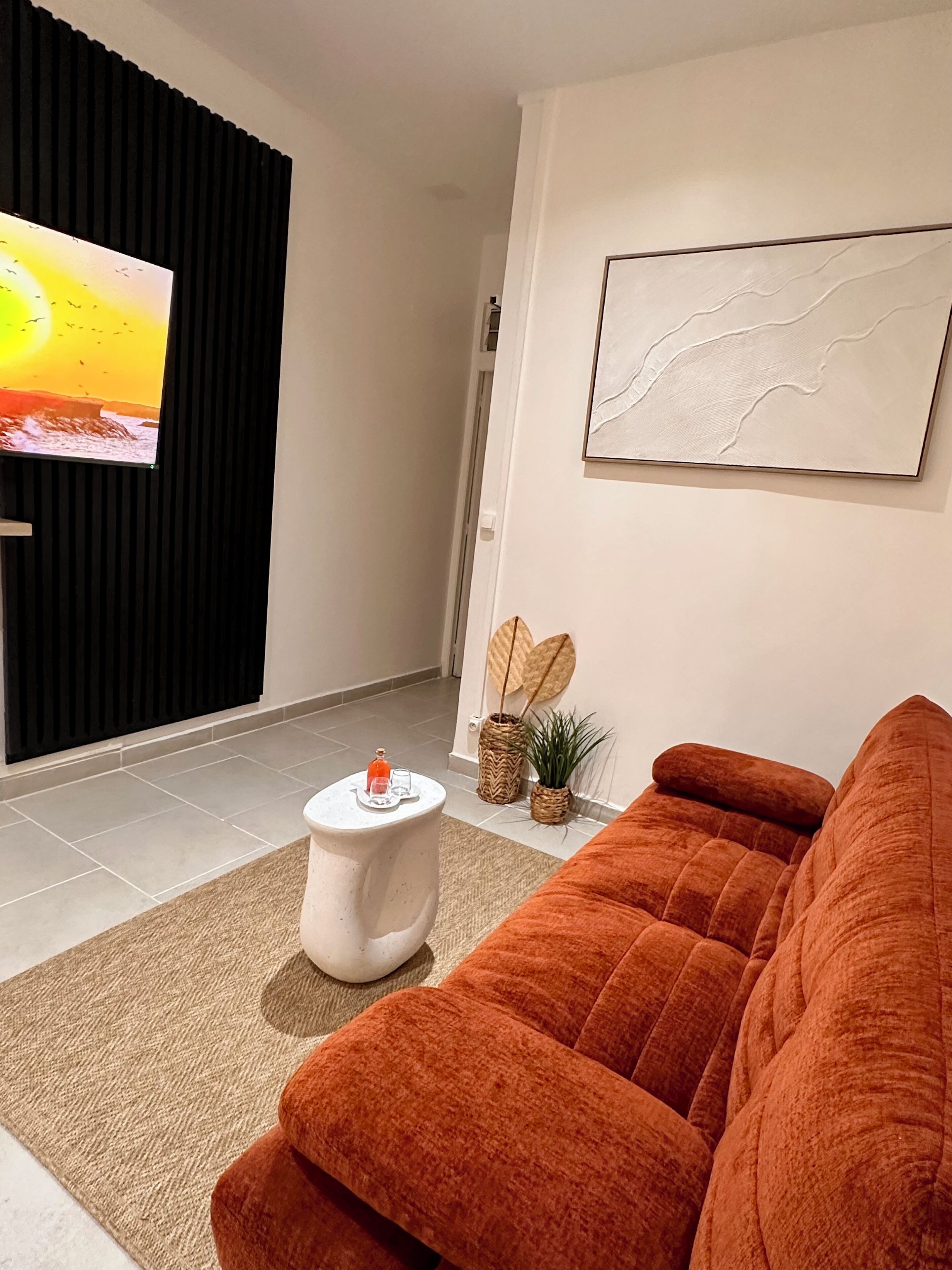 Una sala de estar moderna con un sofá grande color naranja, una mesa de centro blanca, un televisor de pantalla plana montado en un panel negro, una obra de arte minimalista en la pared y una pequeña planta en maceta.