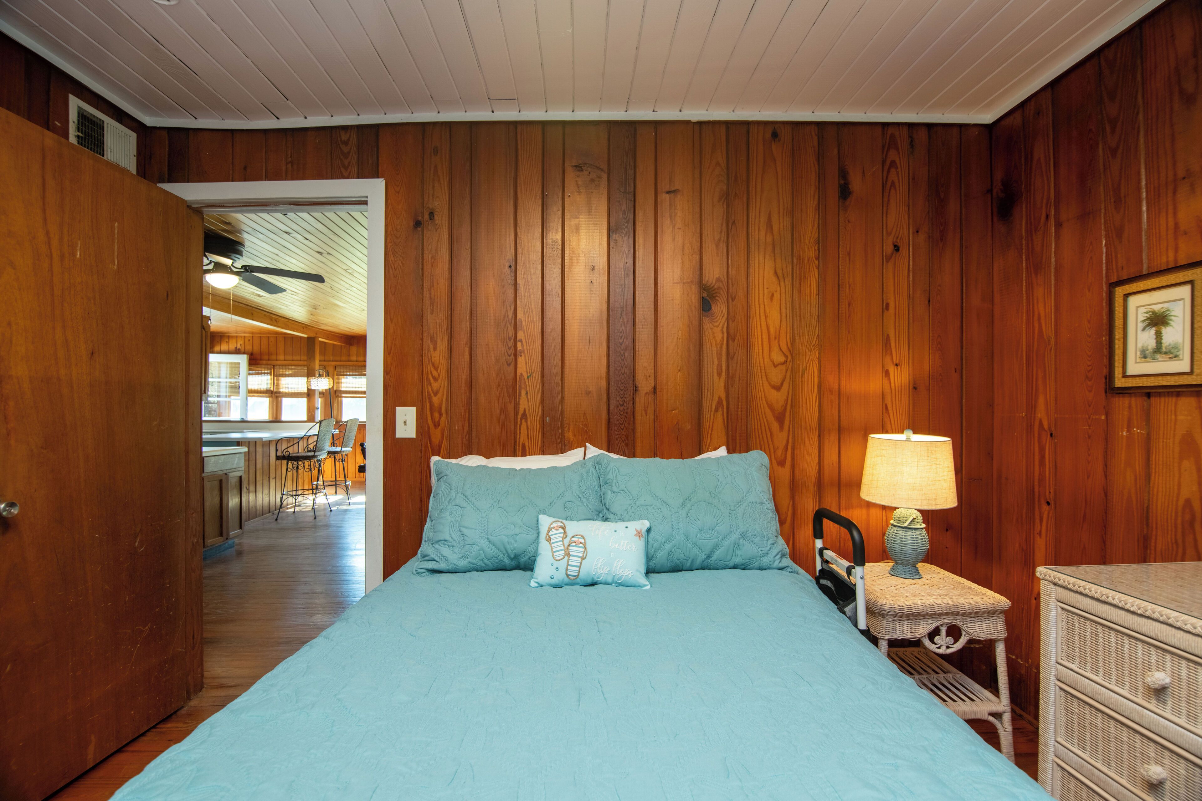 A bedroom with a wooden paneled wall, a bed with blue bedding, a nightstand with a lamp, and a dresser.
