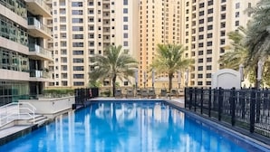 A swimming pool surrounded by tall residential buildings, palm trees, and a fenced area. - Chic Dubai Marina 1BR w Pool and Gym (Dubai)