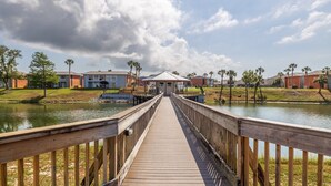A wooden boardwalk leading to a covered pavilion over a body of water, surrounded by residential buildings and palm trees.