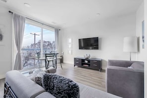 Una sala moderna con un sofá, un televisor montado en la pared, una zona de comedor con mesa y sillas, y un balcón con vistas al exterior.