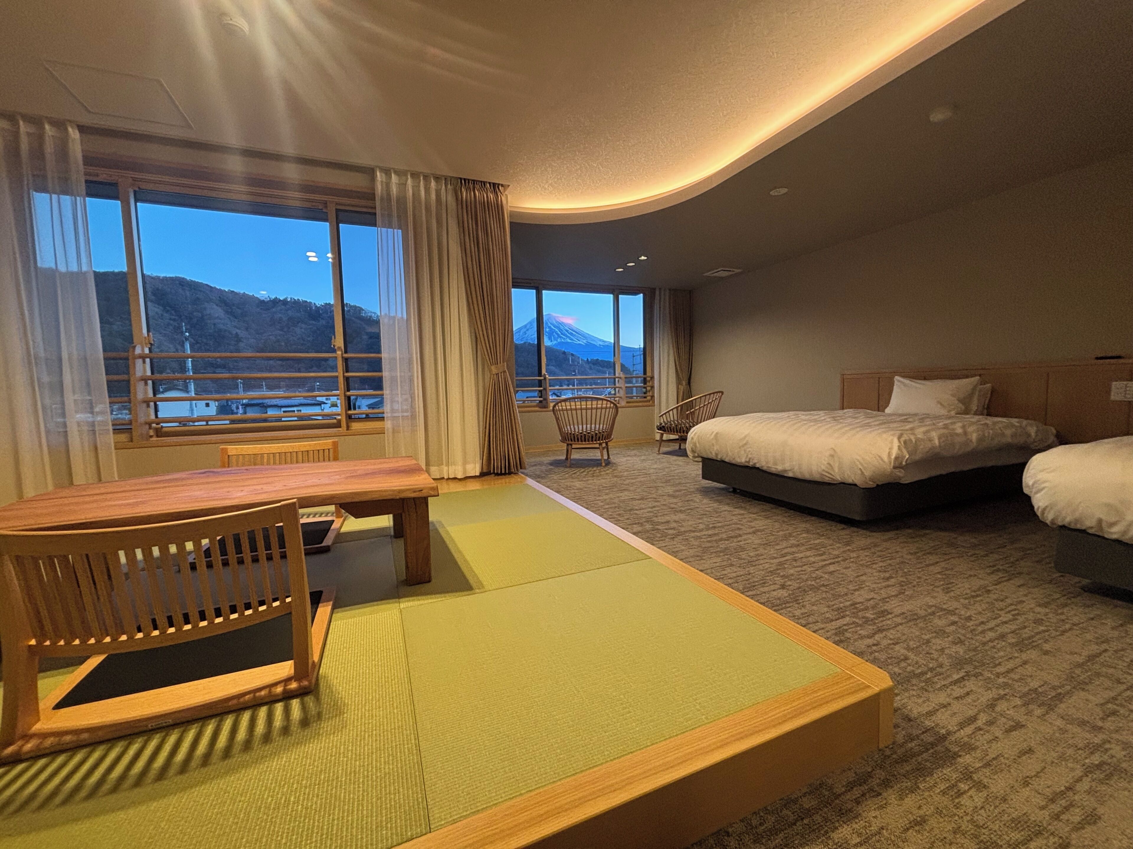 A hotel room with a bed, a table, and chairs, a view of mountains through large windows, and a carpeted floor.