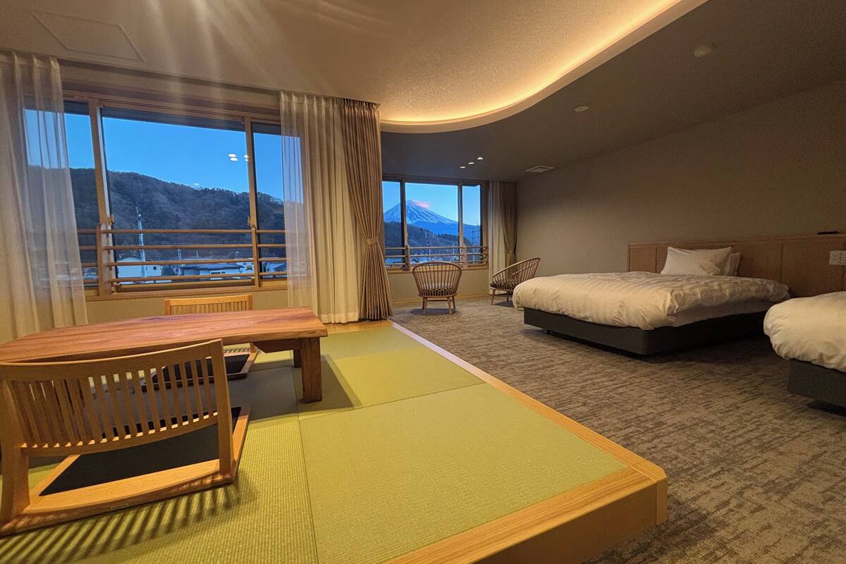 A hotel room with a bed, a table, and chairs, a view of mountains through large windows, and a carpeted floor.