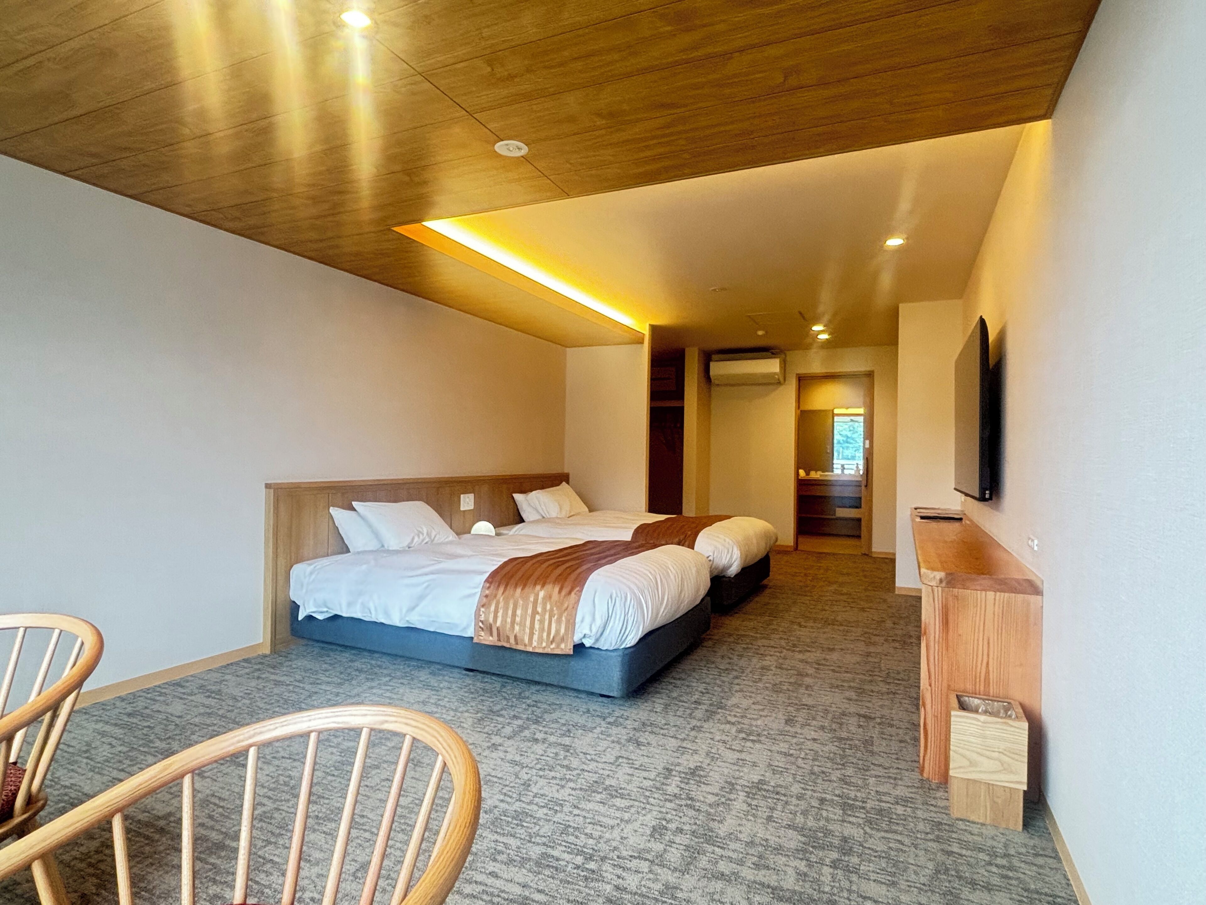 A modern hotel room with a large bed, a wooden headboard, a flat-screen TV mounted on the wall, and a small wooden side table.