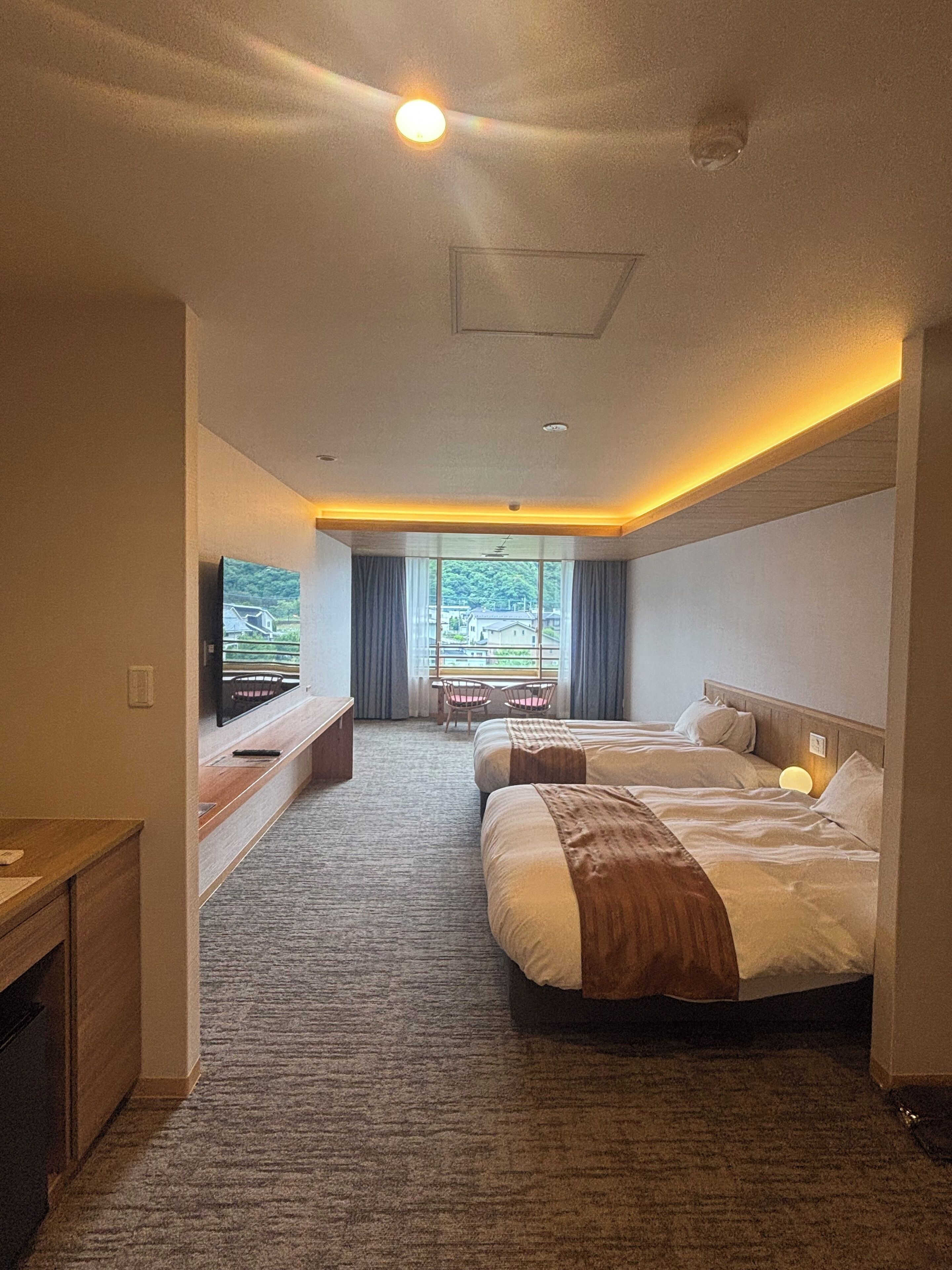 A hotel room with two beds, a TV, a desk, and a view of a green landscape.