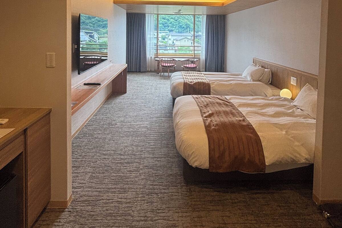 A hotel room with two beds, a TV, a desk, and a window with a view of greenery.