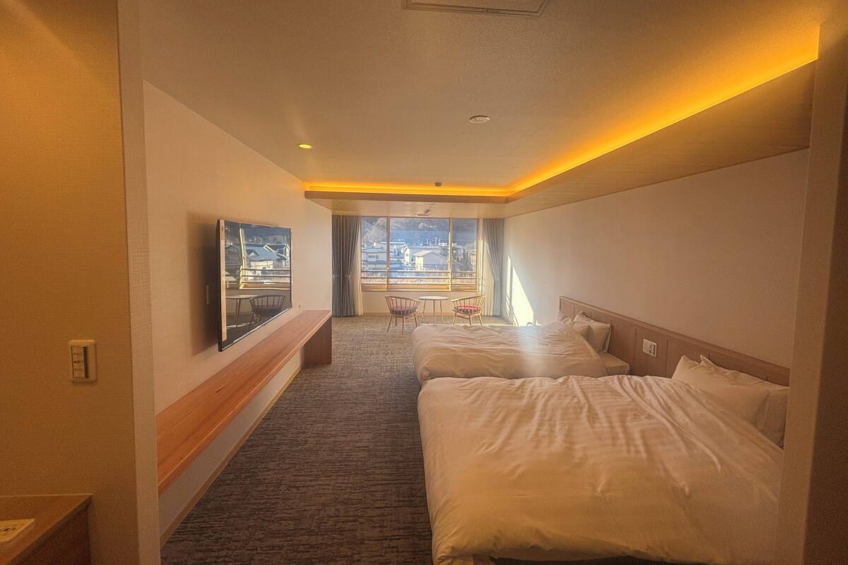 A hotel room with two beds, a TV, a window with a view, and a small table with chairs.