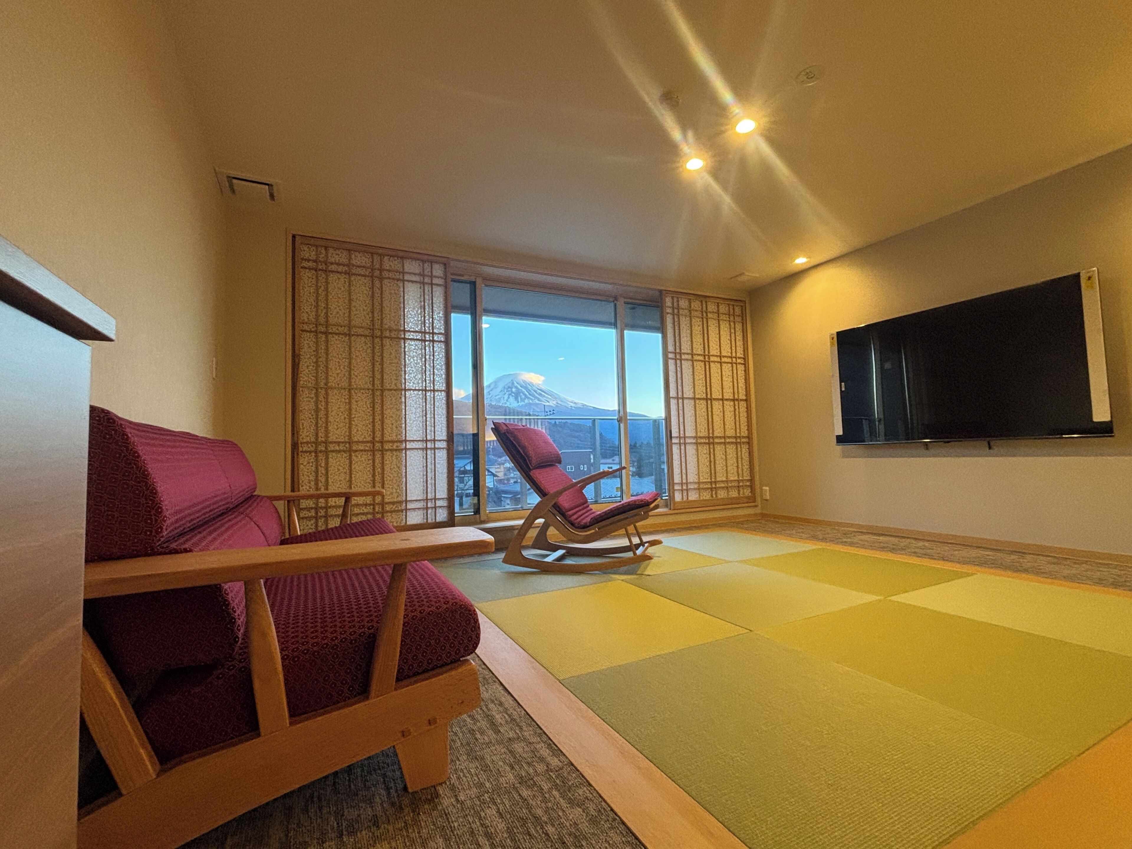 A room with tatami flooring, a tatami mat table, a sofa, a rocking chair, and a TV mounted on the wall.
