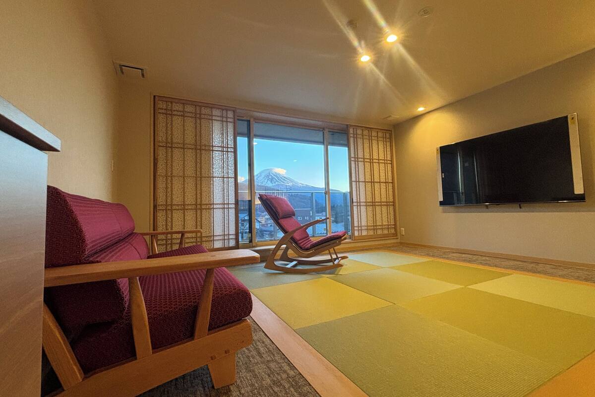 A room with tatami flooring, a tatami mat table, a sofa, a rocking chair, and a TV mounted on the wall.