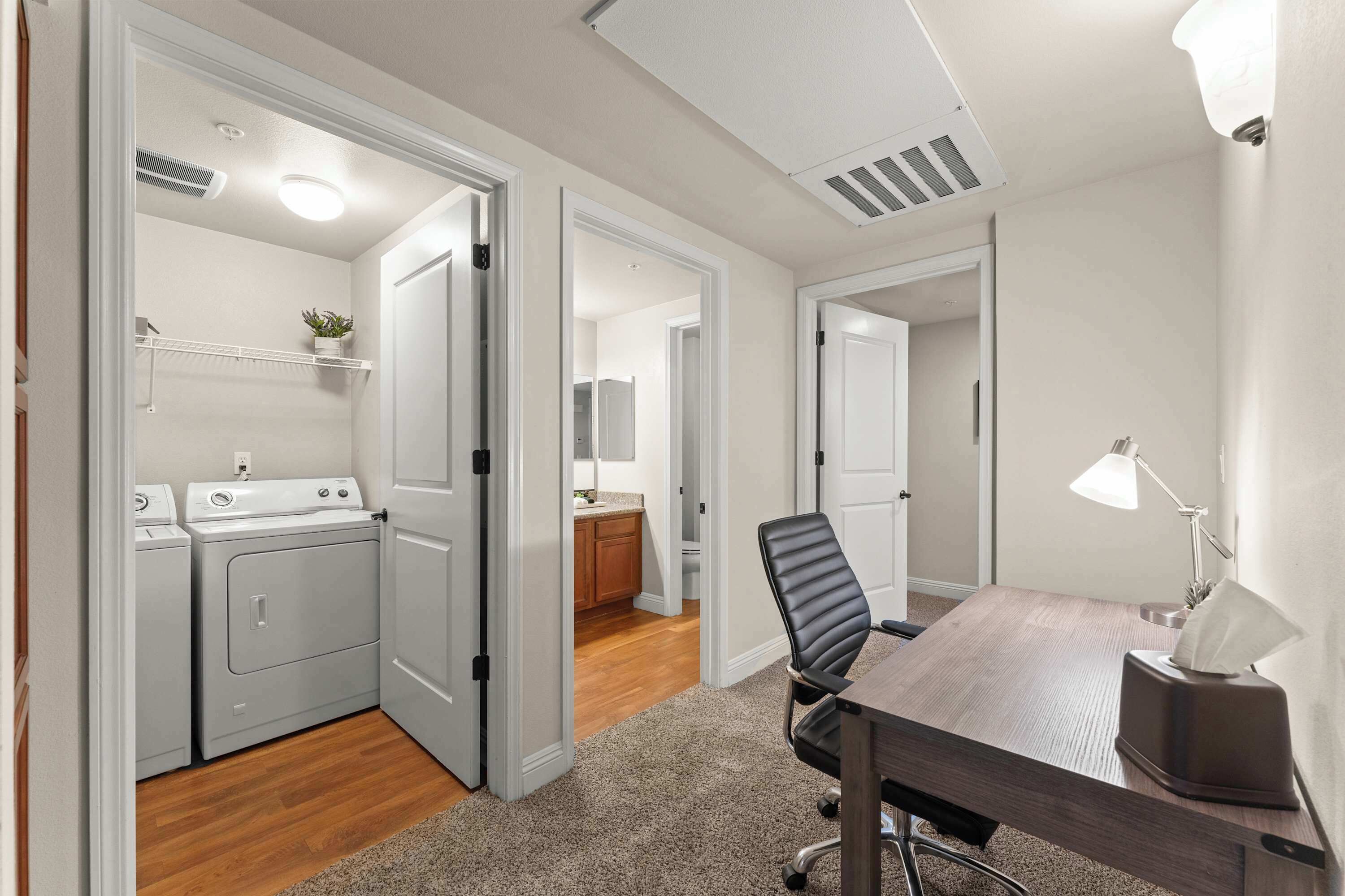A laundry room with a washing machine and dryer, a small plant on a shelf, and a door leading to a kitchen area.