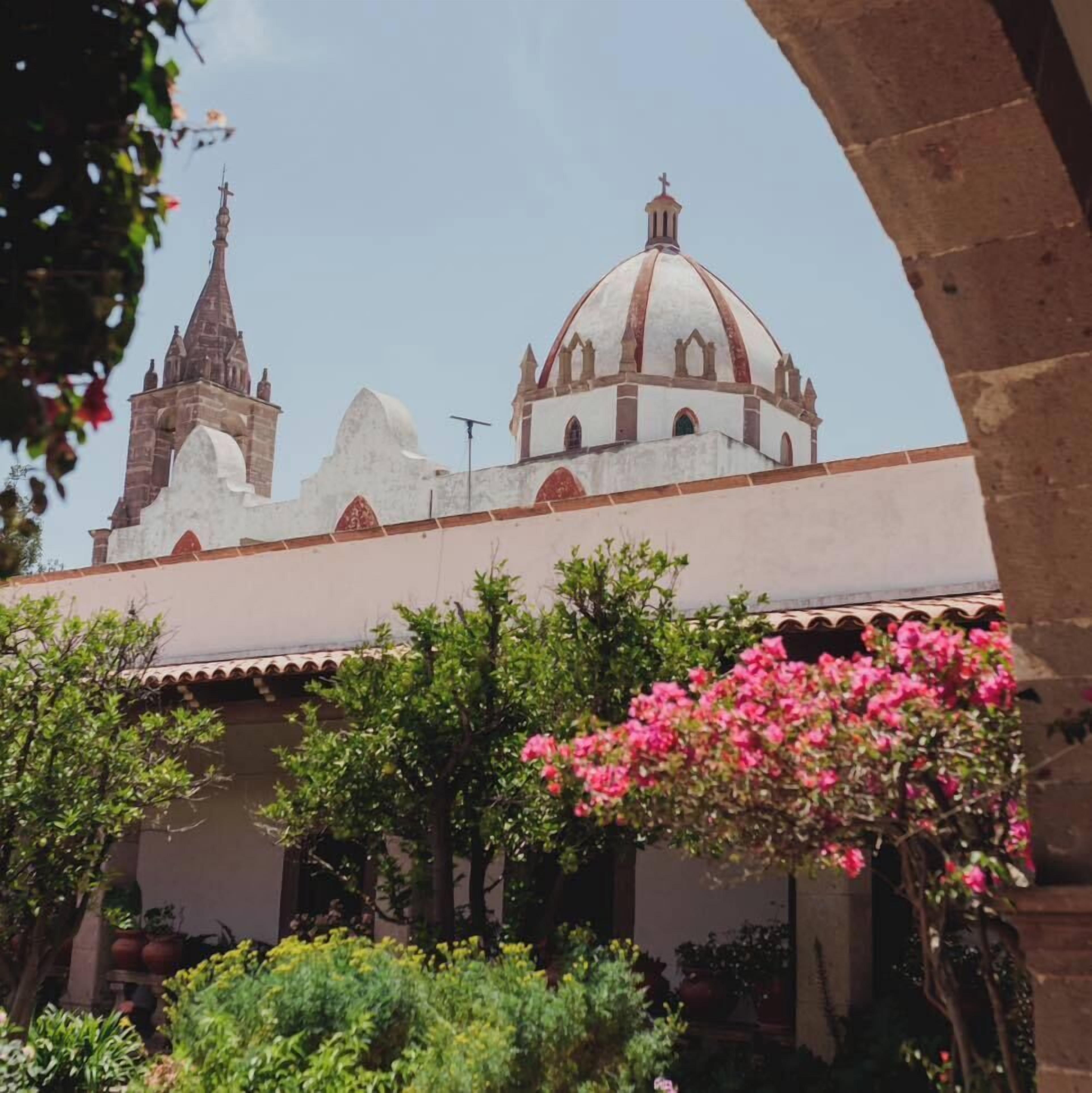 A historical building with a prominent dome and a bell tower, surrounded by lush greenery and flowering plants.