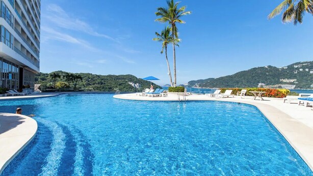 A large, clear swimming pool with lounge chairs and palm trees, surrounded by a modern building and scenic hills in the background. - Charming 2-bedroom apartment in gorgeous Acapulco with AC, WiFi, Ocean View (Acapulco)