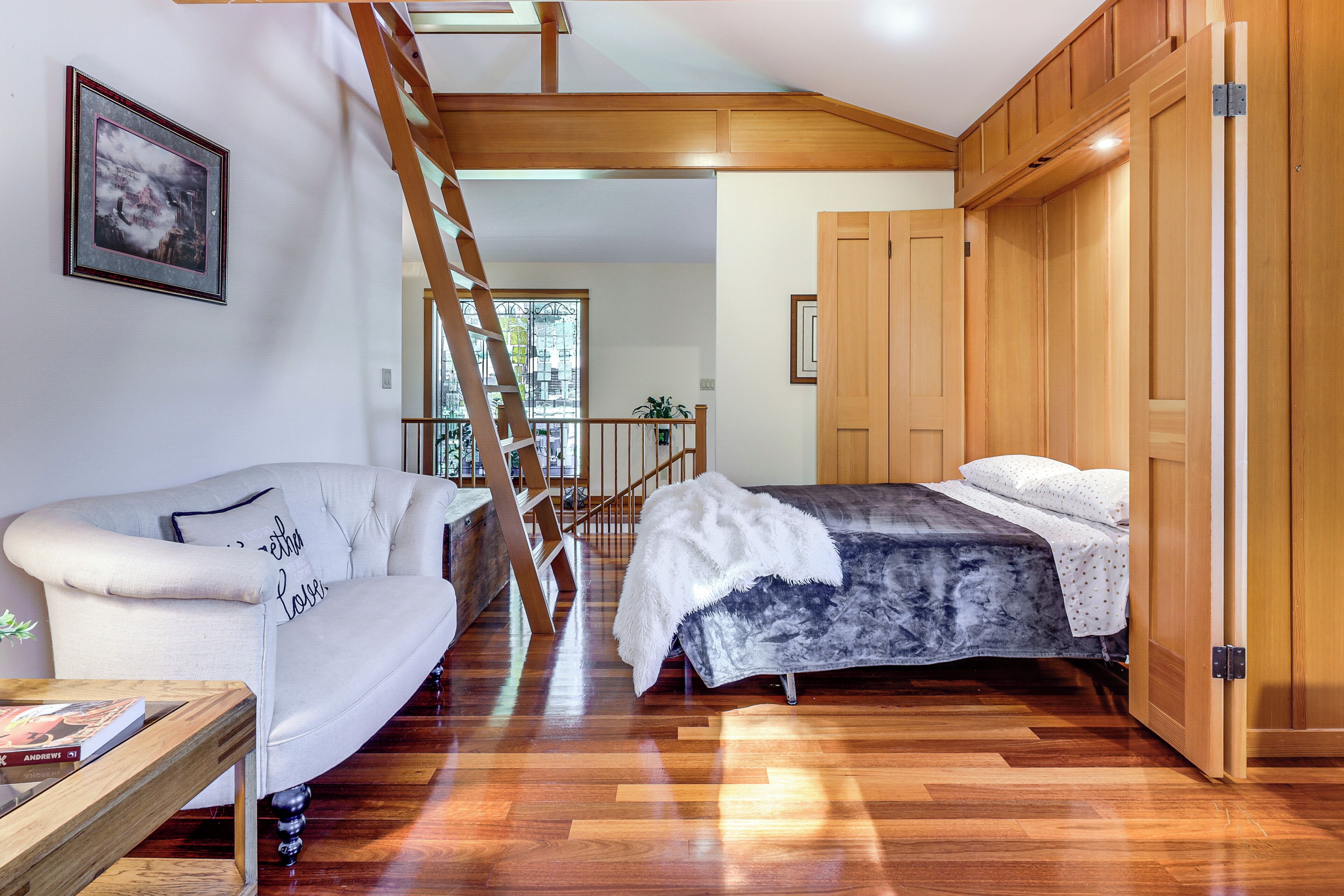 A room with a wooden staircase, a bed with a white and gray bedspread, a white armchair, and a wooden floor.