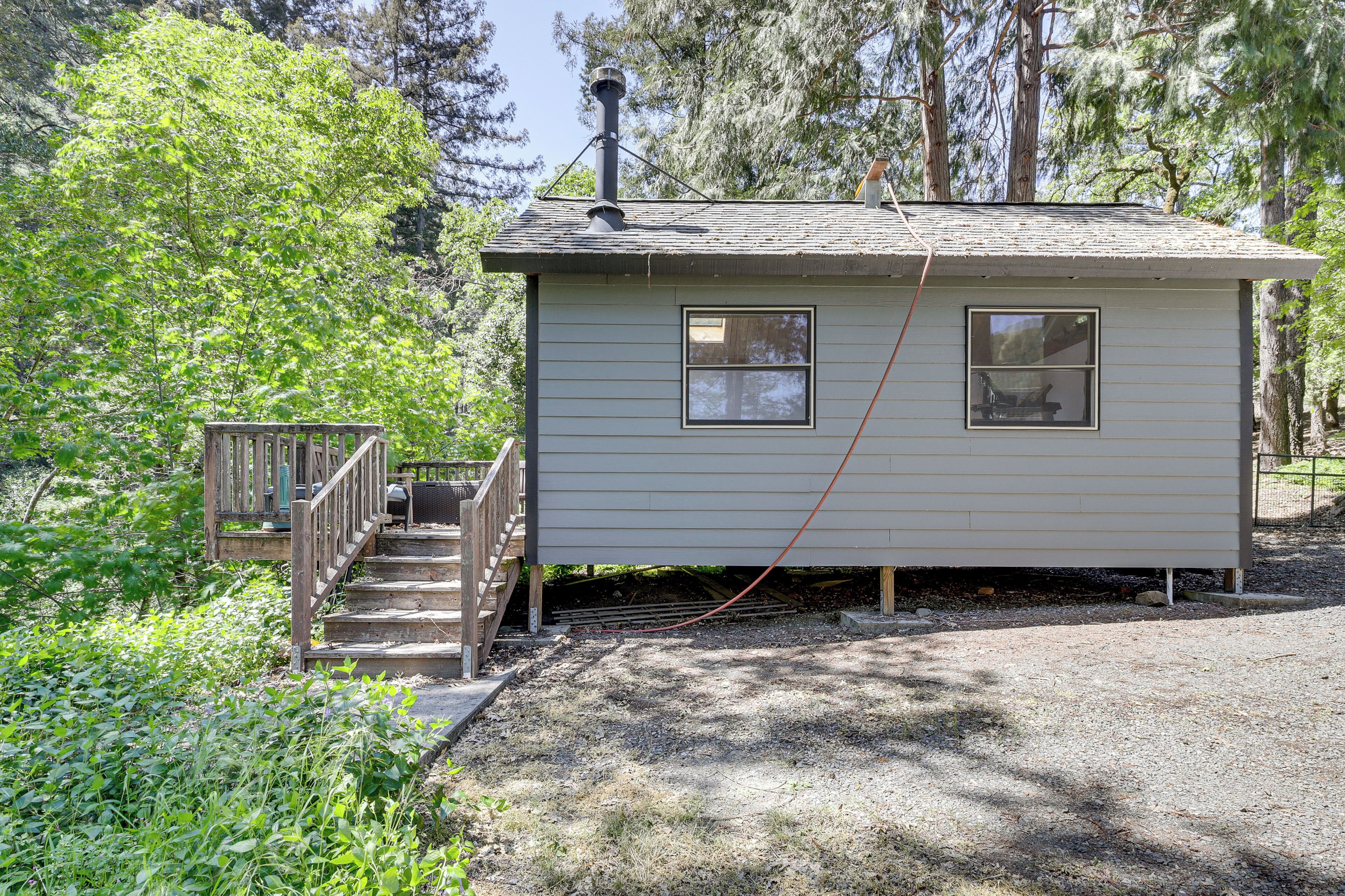 A small, single-story cabin with a wooden deck and a chimney, surrounded by dense trees.
