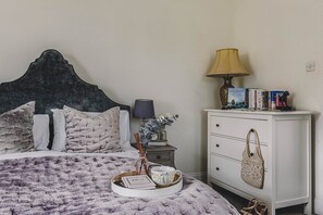 A neatly made bed with a decorative headboard, a bedside table with a lamp, a small shelf with books, and a drawer unit with a woven bag.