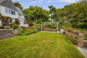 A house with a white exterior, a stone wall, a wooden bench, and a pergola in a garden.