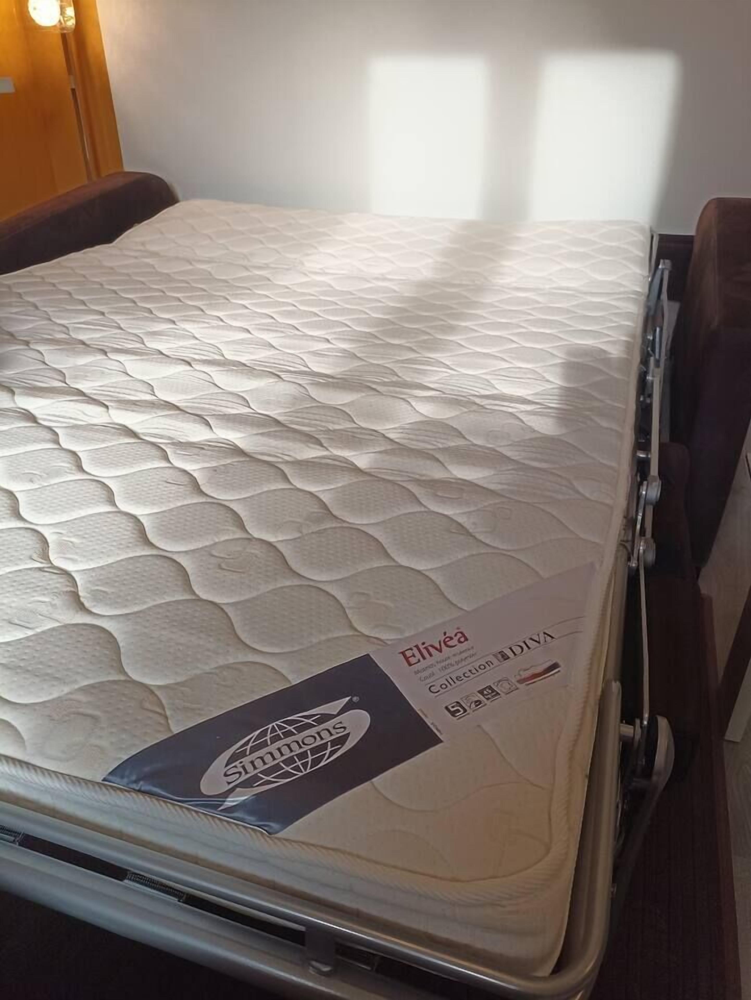 A mattress with a branded label, a wooden headboard, and a wall with a window.