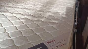 A mattress with a branded label, a wooden headboard, and a wall with a window.