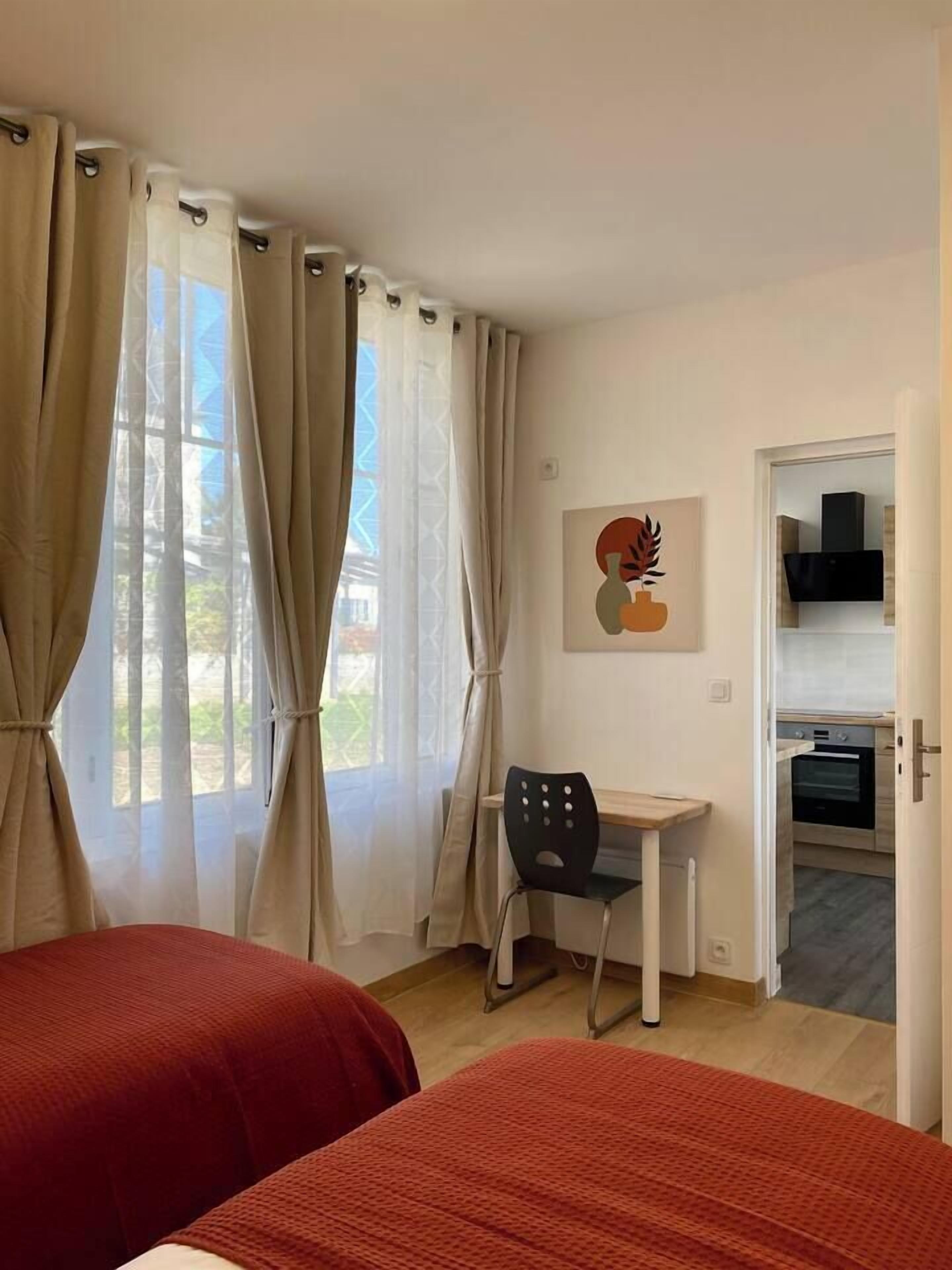 A room with two beds, a desk, a chair, a painting, and a kitchenette.