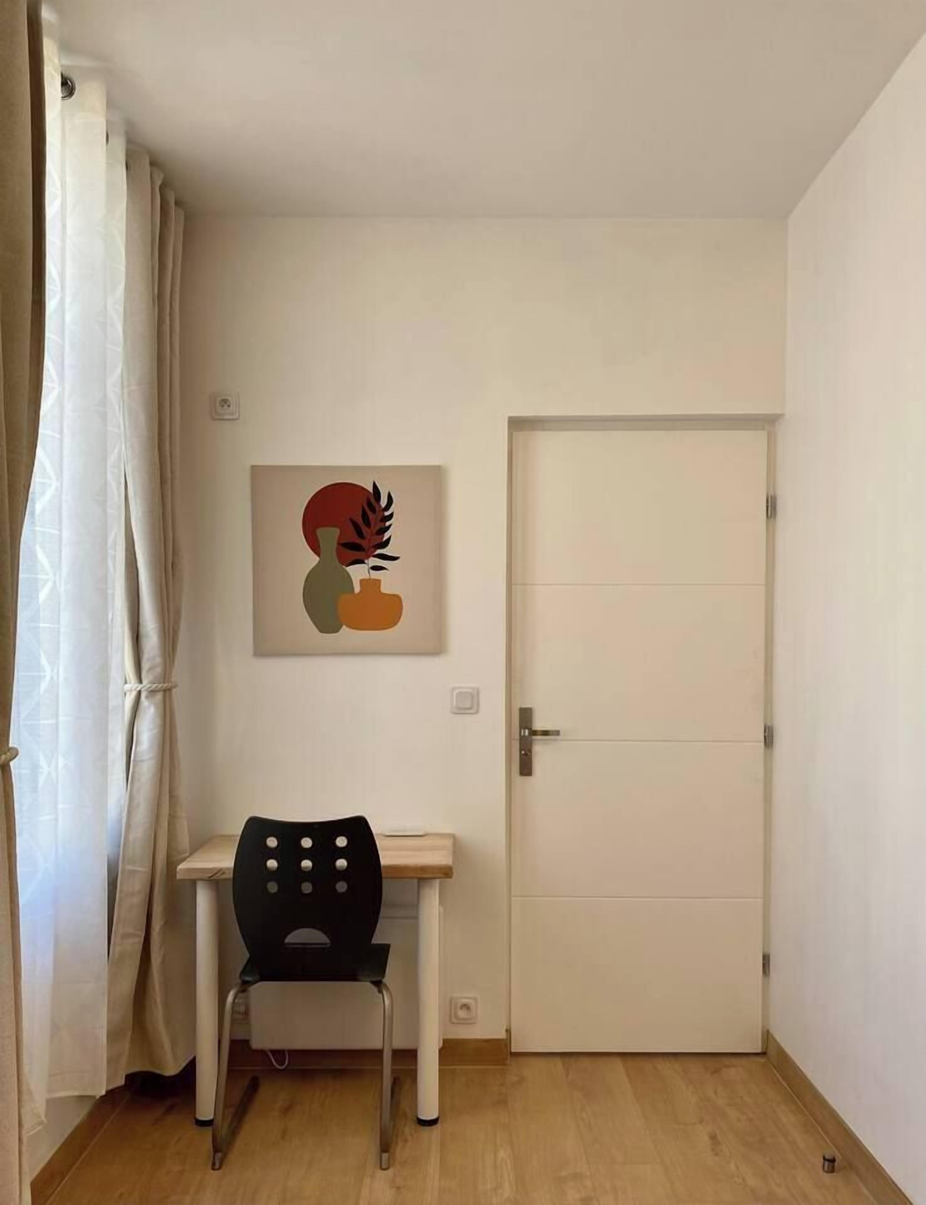 Ett litet rum med en vit dörr, en svart stol, ett litet bord och ett abstrakt konstverk som hänger på väggen.