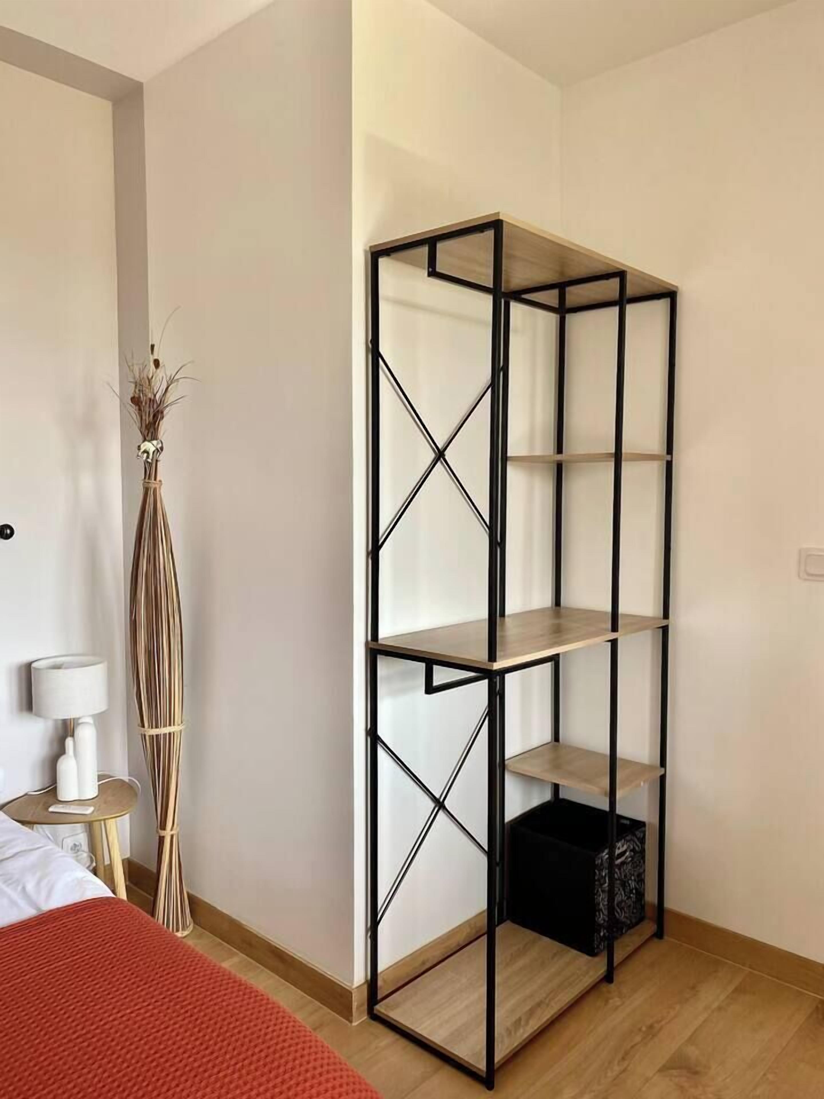 A room with a tall, black metal shelving unit with three shelves, a bedside table with a lamp, and a red bedspread.