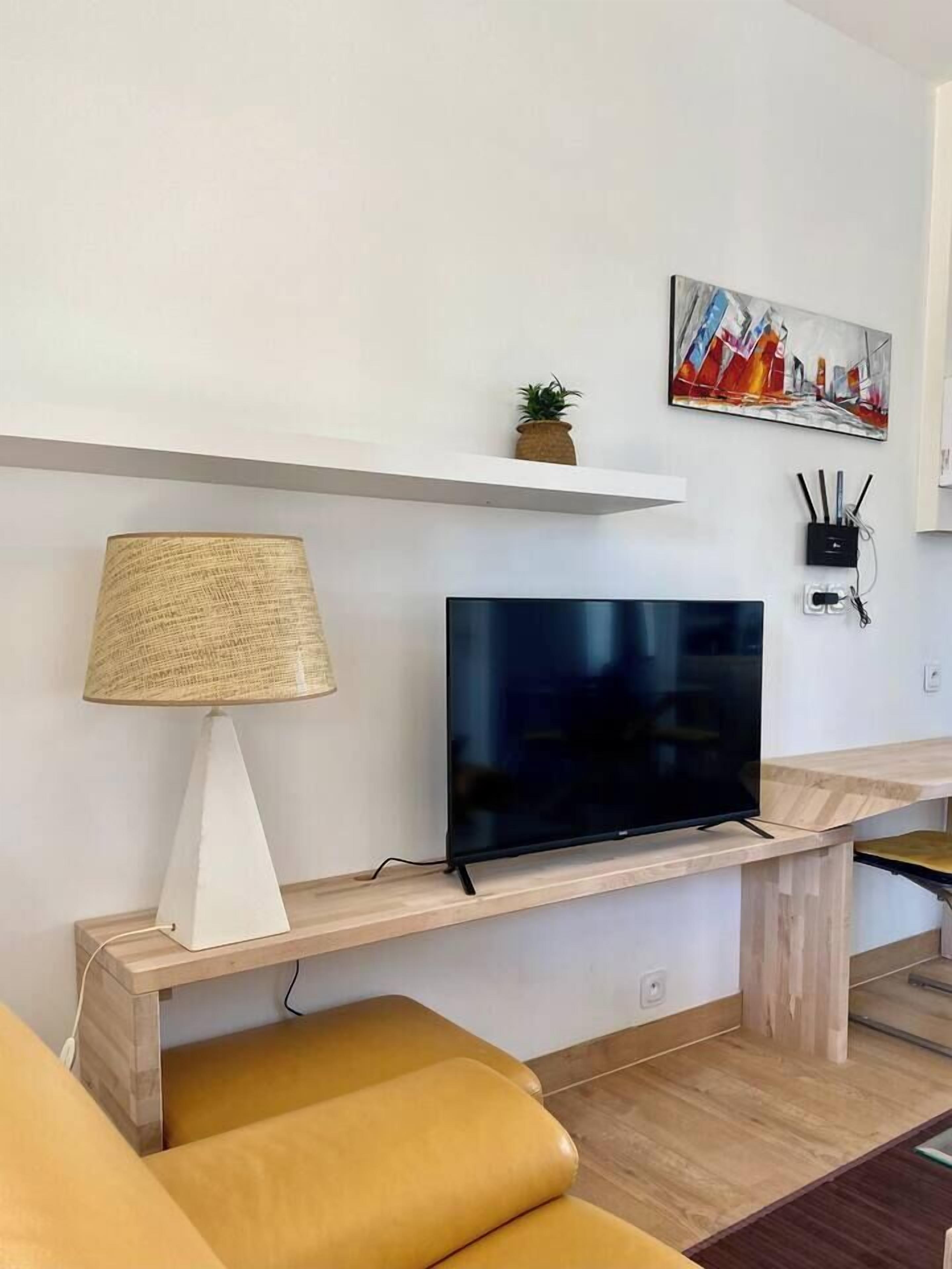 Ein modernes Wohnzimmer mit Fernseher auf einem Holzständer, einer Lampe, einer kleinen Pflanze und einem Gemälde an der Wand.