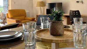 Un coin repas avec une table ronde dressée pour deux personnes, un canapé, un téléviseur et un tableau accroché au mur.
