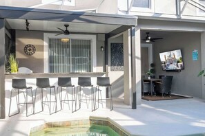 Un espacio al aire libre cubierto con bar, sillas, piscina y televisor.