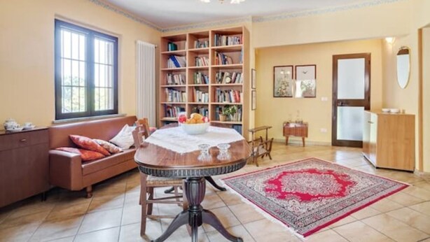A cozy living room with a round wooden table, a bookshelf, a sofa, and a large window.