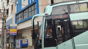 Ein hellblauer Bus mit der Aufschrift 'WELCOME' auf der Vorderseite, geparkt vor einem Gebäude mit der Beschilderung 'HOTEL SWAGATH GRAND ROOMS & FUNCTION HALL'.