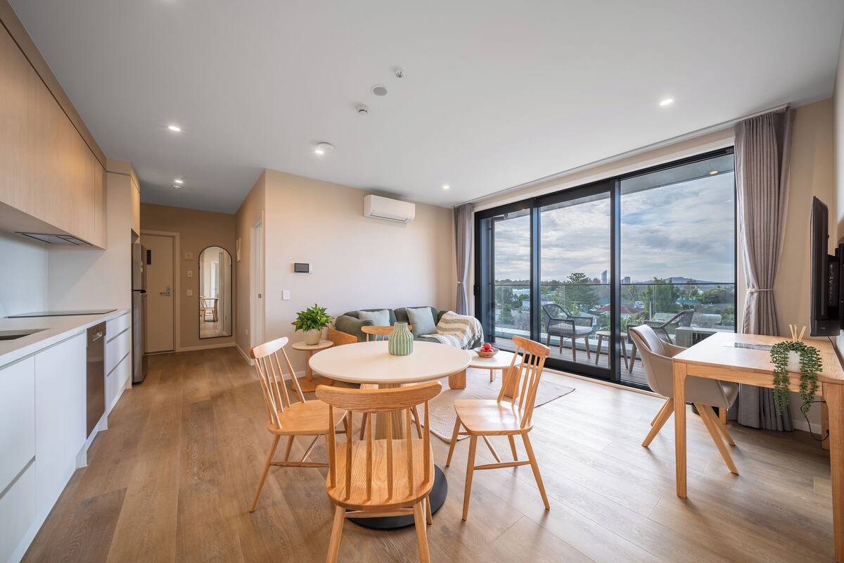 A modern kitchen with wooden cabinets, a dining area with a round table and chairs, and a large window offering a city view.