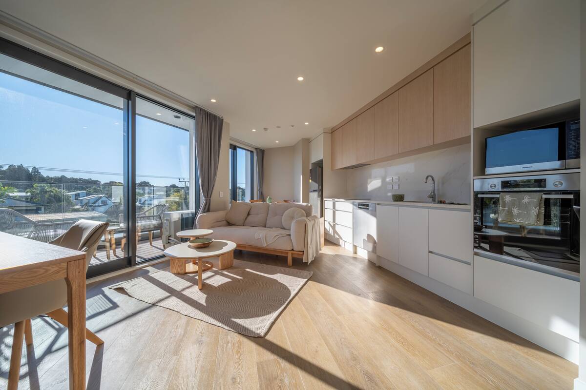 A modern kitchen with built-in appliances, a dining area with a wooden table and chairs, and a large sliding glass door leading to a balcony with outdoor furniture.