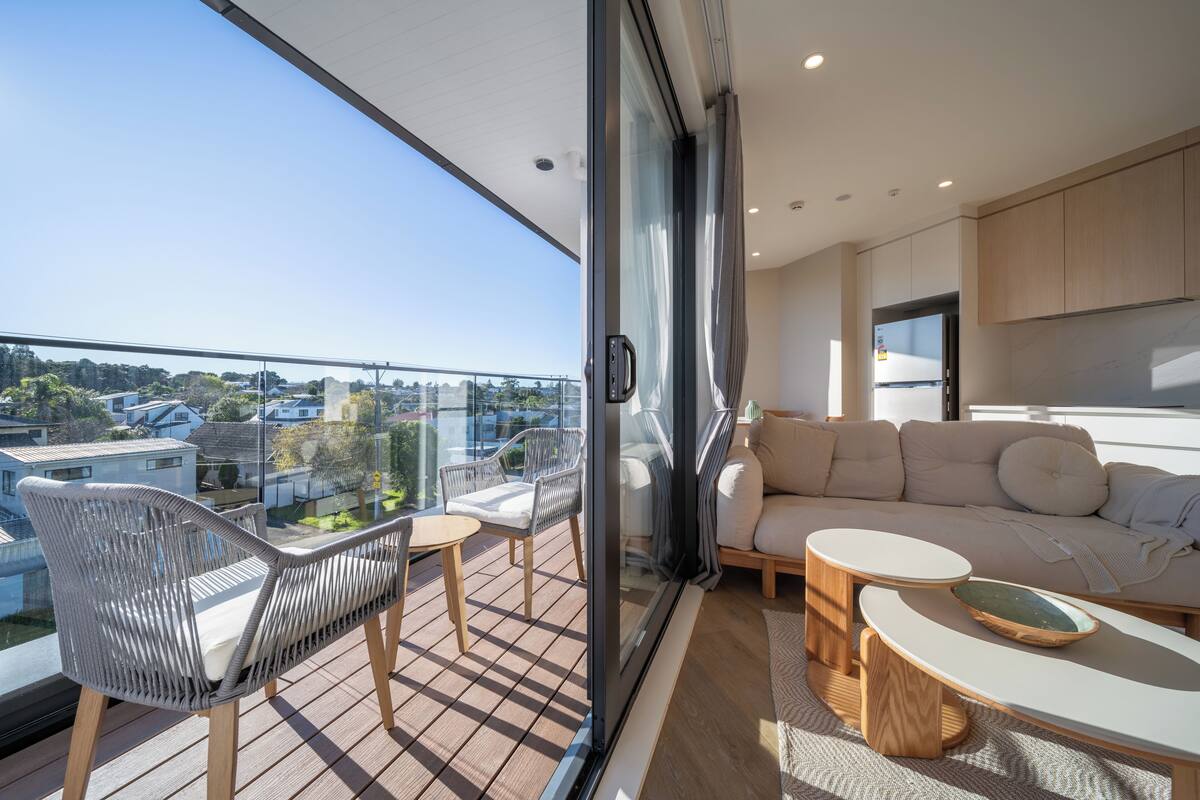 A modern living room with a sofa, chairs, and a small table. A large glass door leads to a balcony with a view of the city.