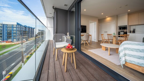 Uma varanda moderna com piso de madeira, área de refeições com mesa e cadeiras, cama com manta listrada e cozinha com armários e eletrodomésticos.