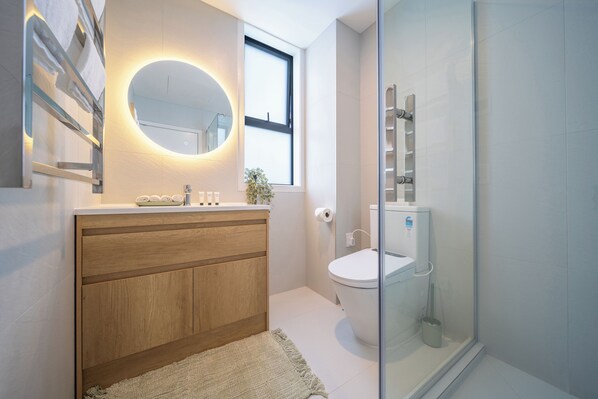 A modern bathroom with a wooden vanity, a round mirror, a toilet, and a glass shower enclosure. - Palm Suites - Auckland (Auckland)