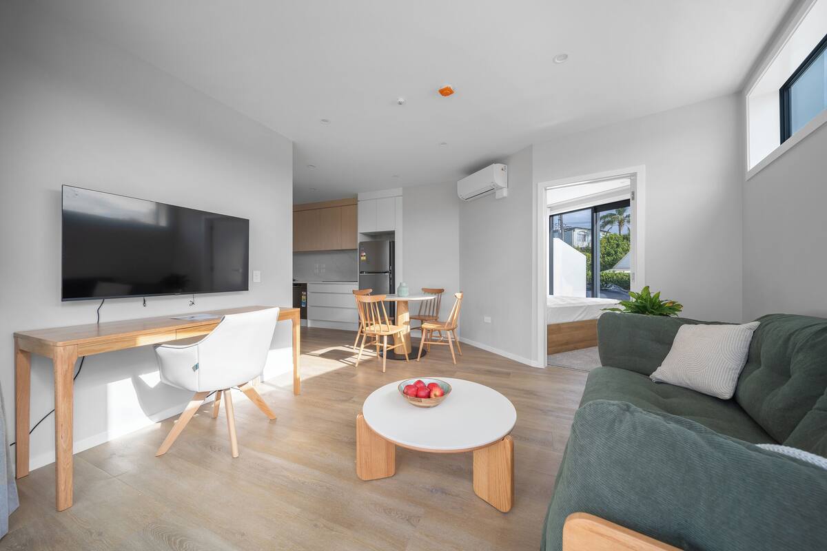 A modern living space with a flat-screen TV, wooden desk, white chair, round dining table, and a green sofa with a white pillow.