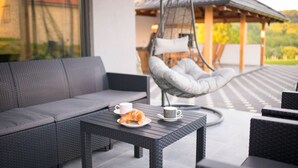 A modern outdoor seating area with a grey sofa, a wicker table, a hanging chair, and a croissant on the table. - Villa Old Hills by Villas Guide (Ljubešcica)